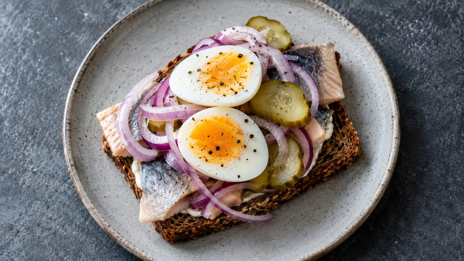 Smørrebrød på rugbrød med sild, hårdkogt æg, rødløg og syltet agurk